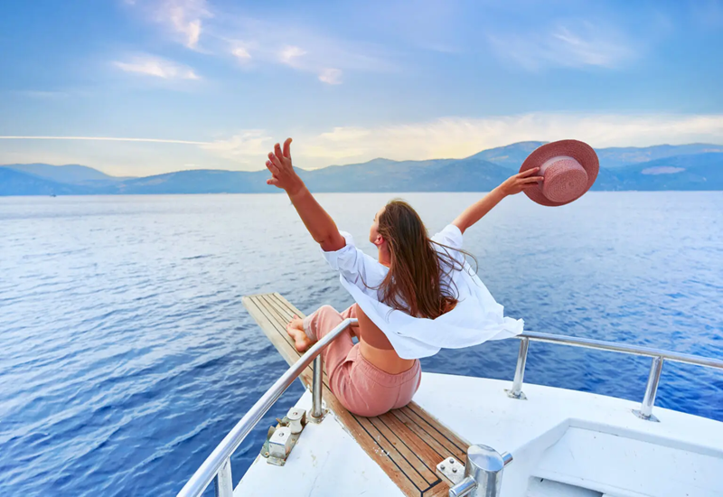 Woman enjoying a boat ride with arms raised and hat in hand.